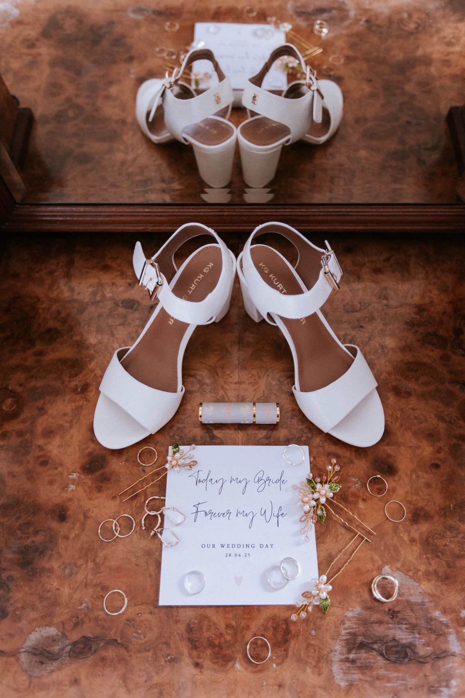 White bridal sandals and wedding rings displayed with a note on brown surface.