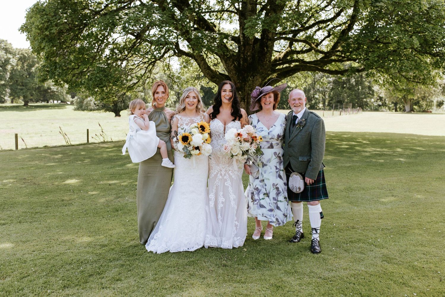 Wedding party members gather for formal photos on a sunny lawn with trees in background.