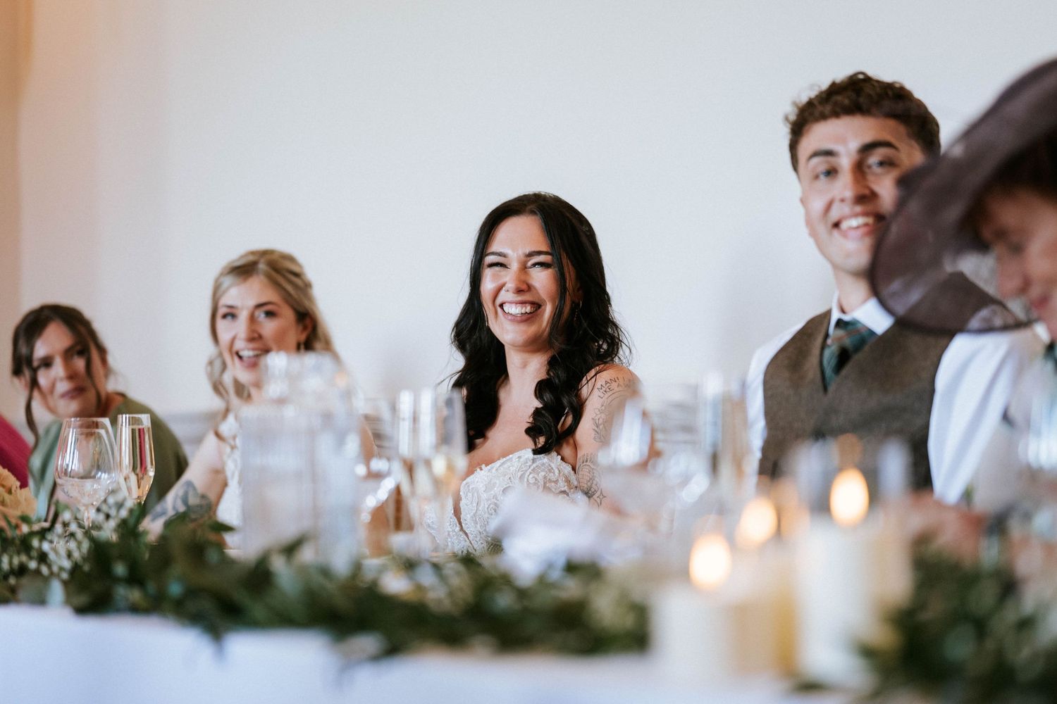 Wedding guests laugh and smile during dinner reception with candlelit table and greenery decorations.