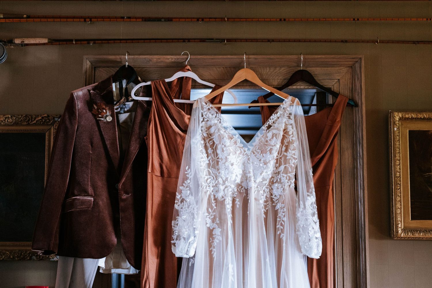 Wedding attire including white lace dresses and brown suits hanging on wooden hangers in preparation for the ceremony.