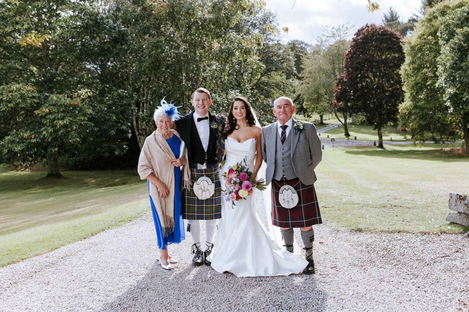 Small wedding group in Scottish formal wear poses on gravel path surrounded by green gardens.