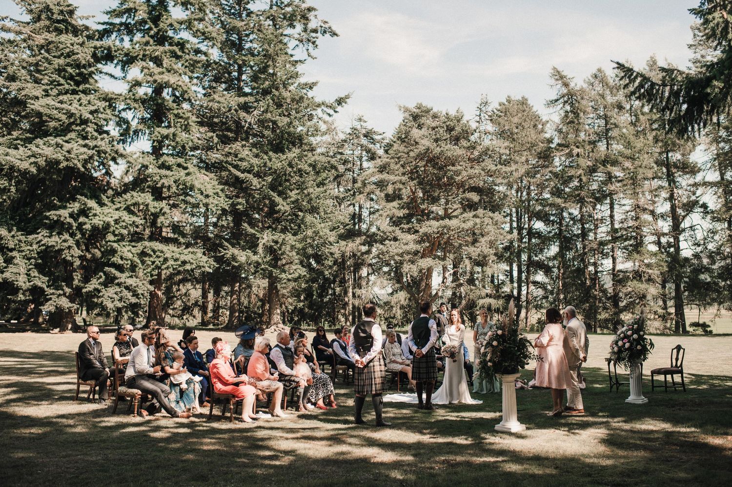 Wide shot of wedding ceremony setup in wooded outdoor setting with guests seated.