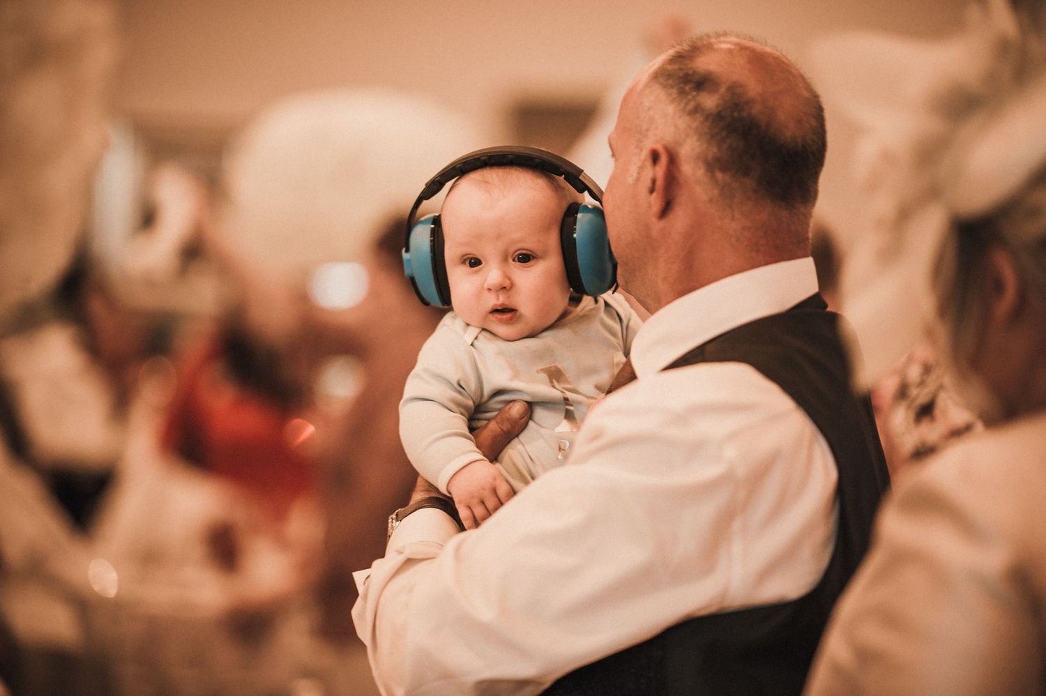Baby wearing protective headphones while being held at a formal event.