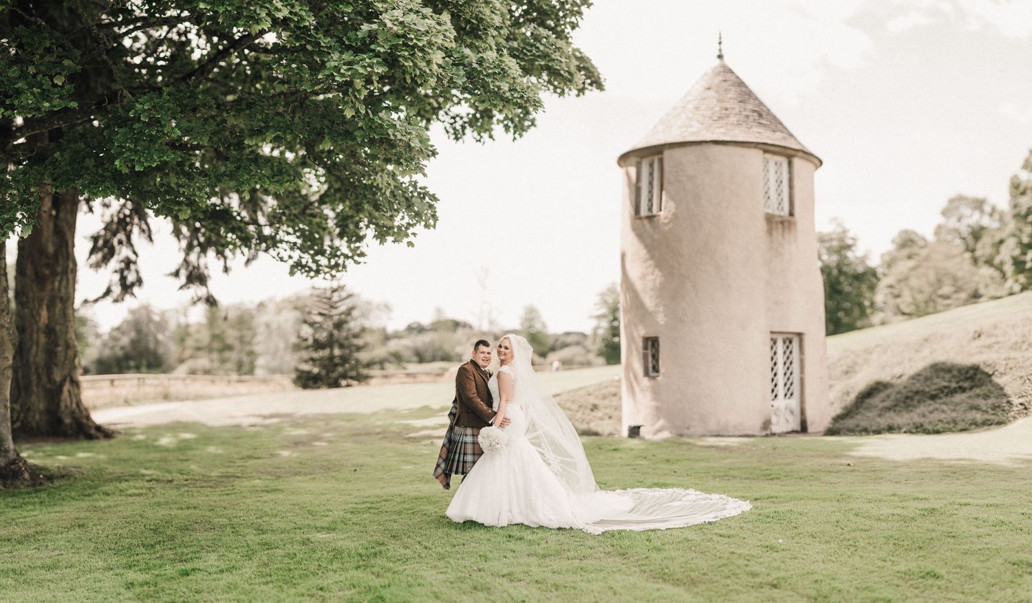 Wedding couple poses near historic stone tower on elegant garden grounds.