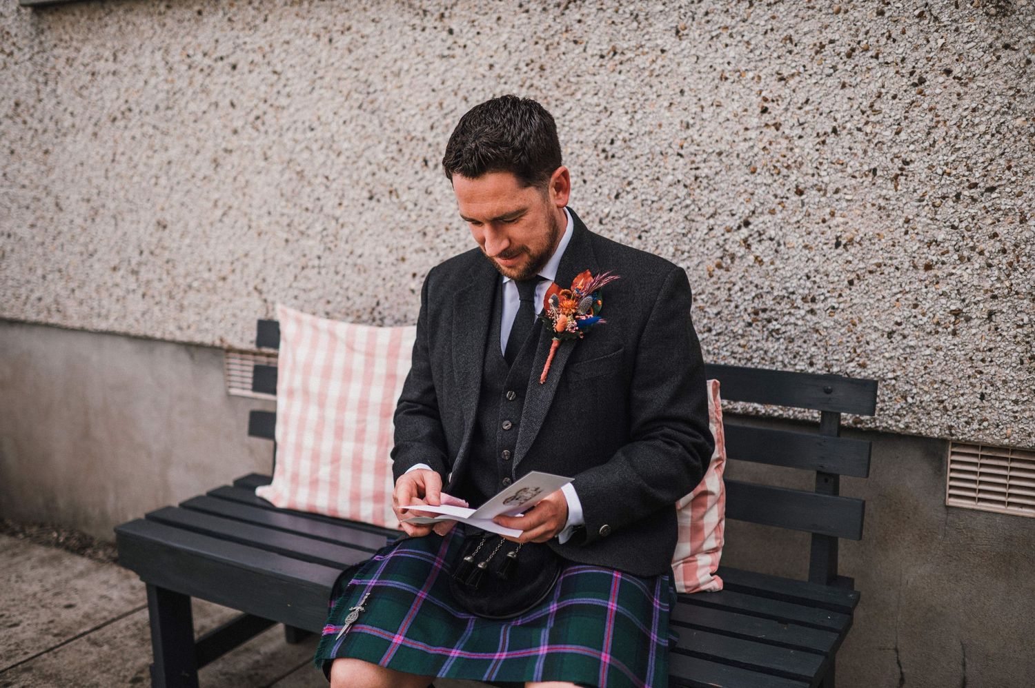 Close-up of someone in formal Scottish attire sitting on a bench with pink cushion.