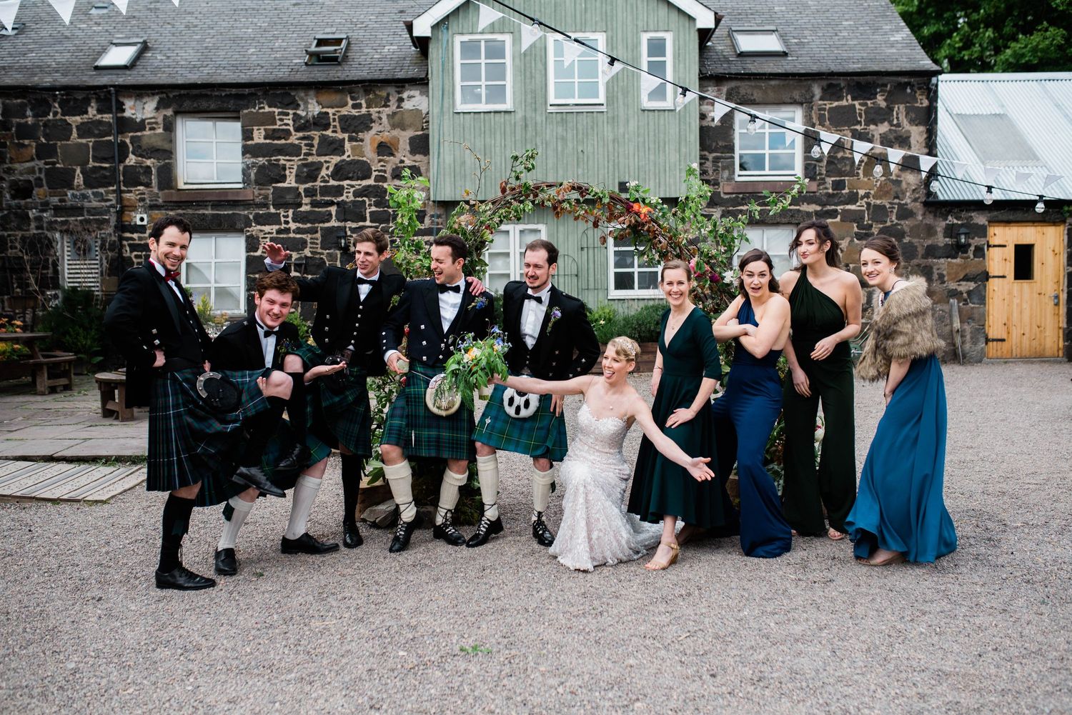 Wedding party in formal attire and kilts pose casually on gravel outside a stone building.