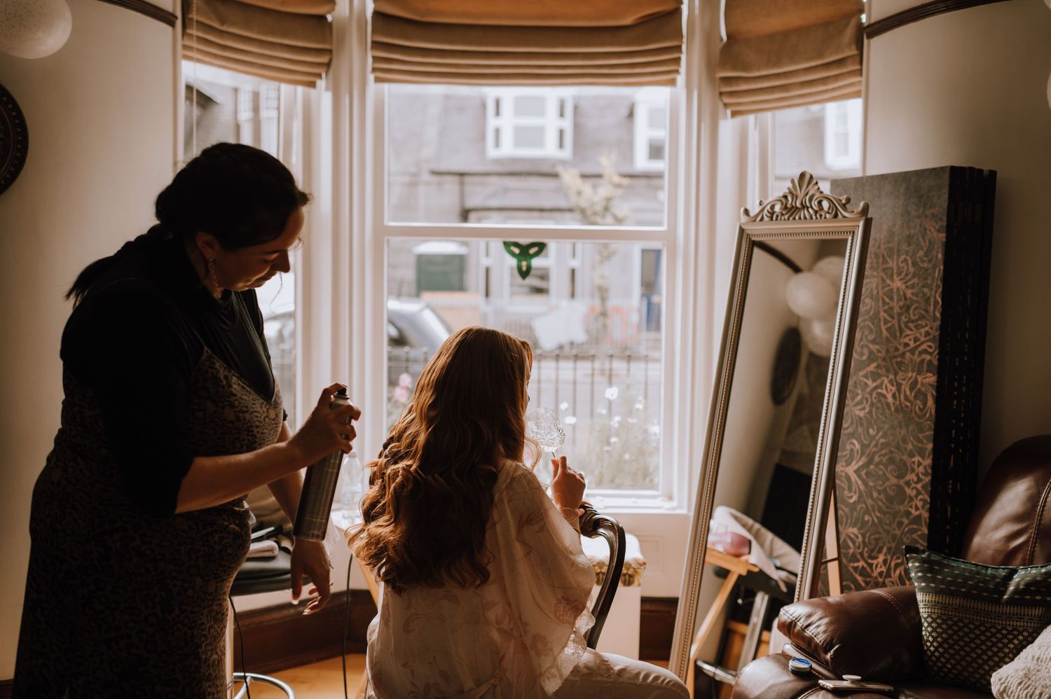 Getting ready scene near a bay window with hair and makeup preparation in a bright room.