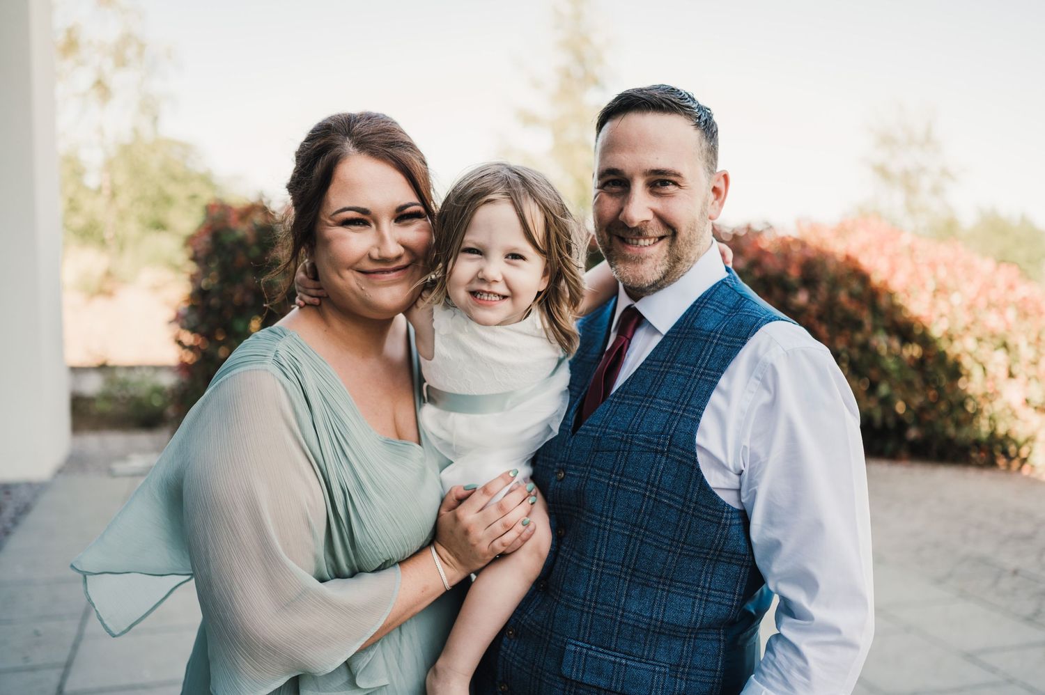 Family portrait taken outdoors during sunny day with foliage backdrop.