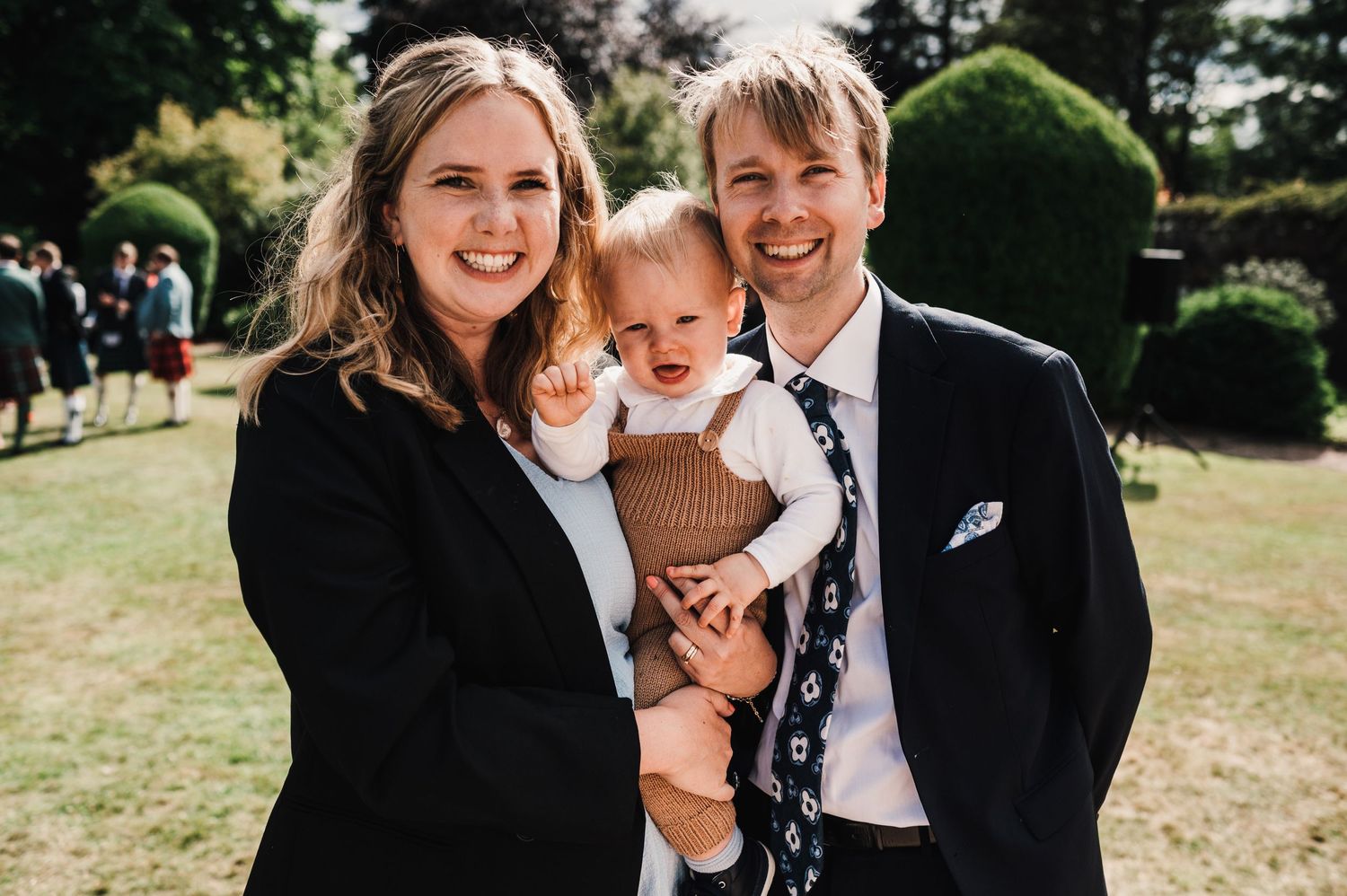 Family poses together outdoors on a sunny day with a small baby in a brown sweater.