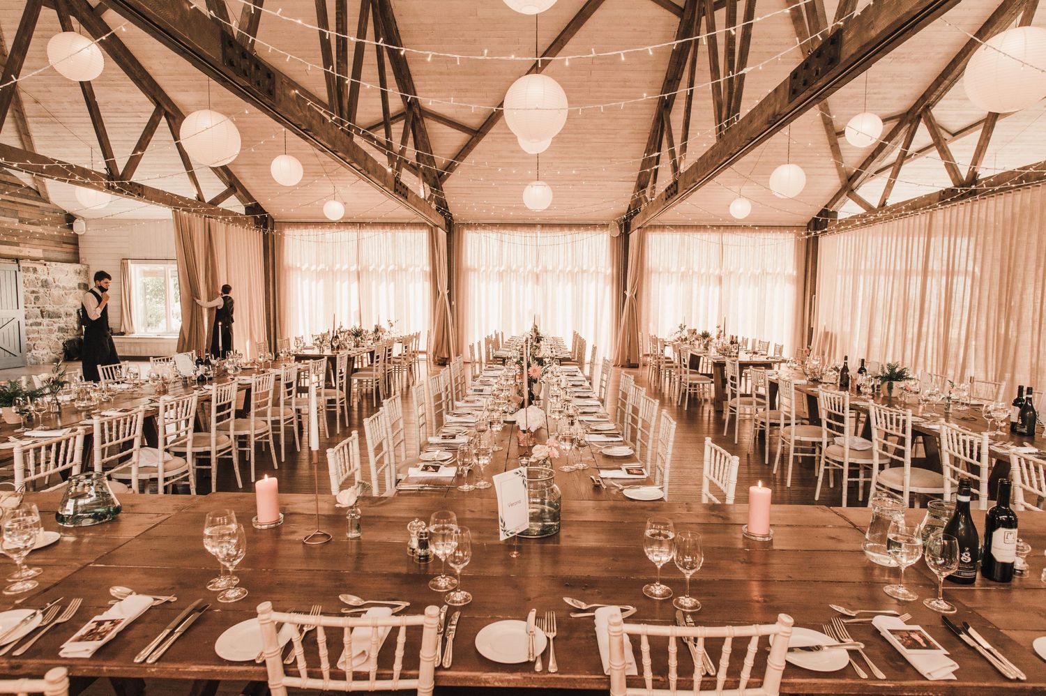 Rustic barn wedding reception hall set with long wooden tables and white chairs under exposed beams.
