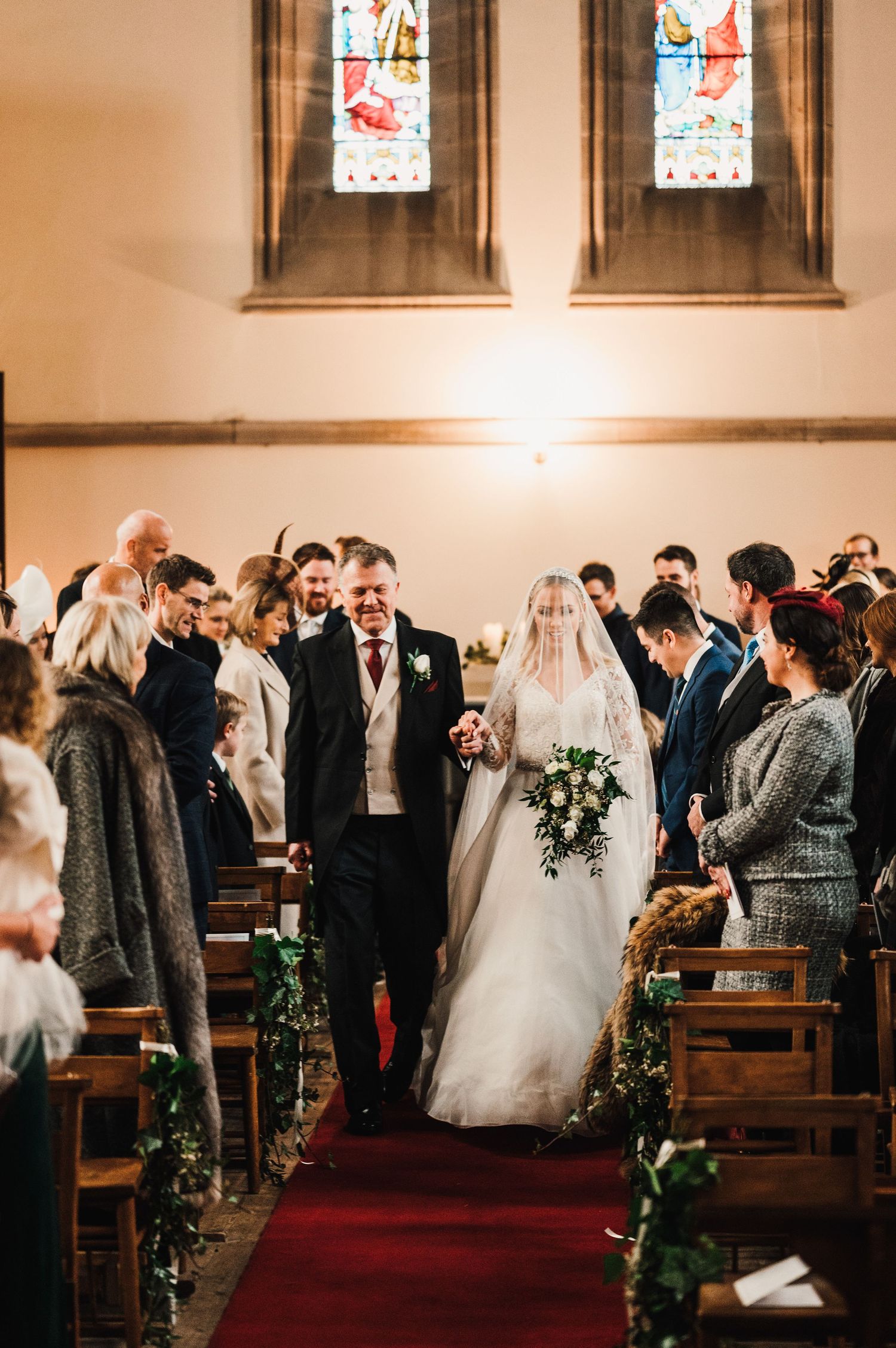 A traditional church wedding ceremony with guests watching the procession down a red-carpeted aisle.