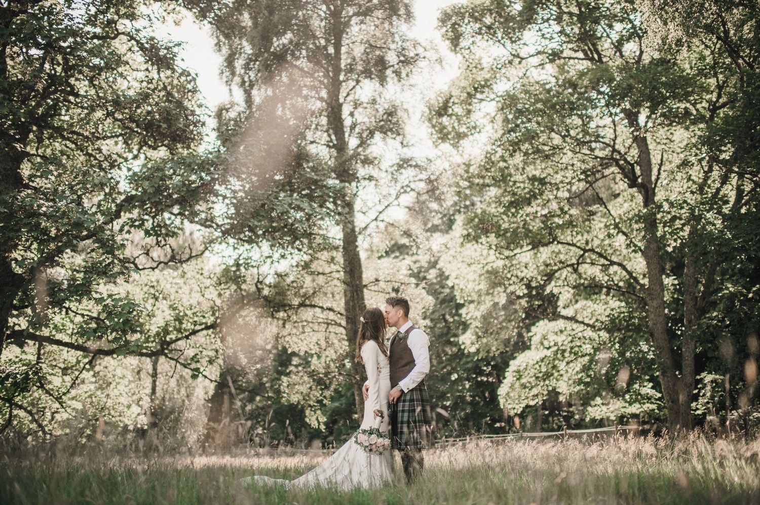 A wedding couple shares an intimate moment in a scenic woodland setting with natural sunlight filtering through trees.