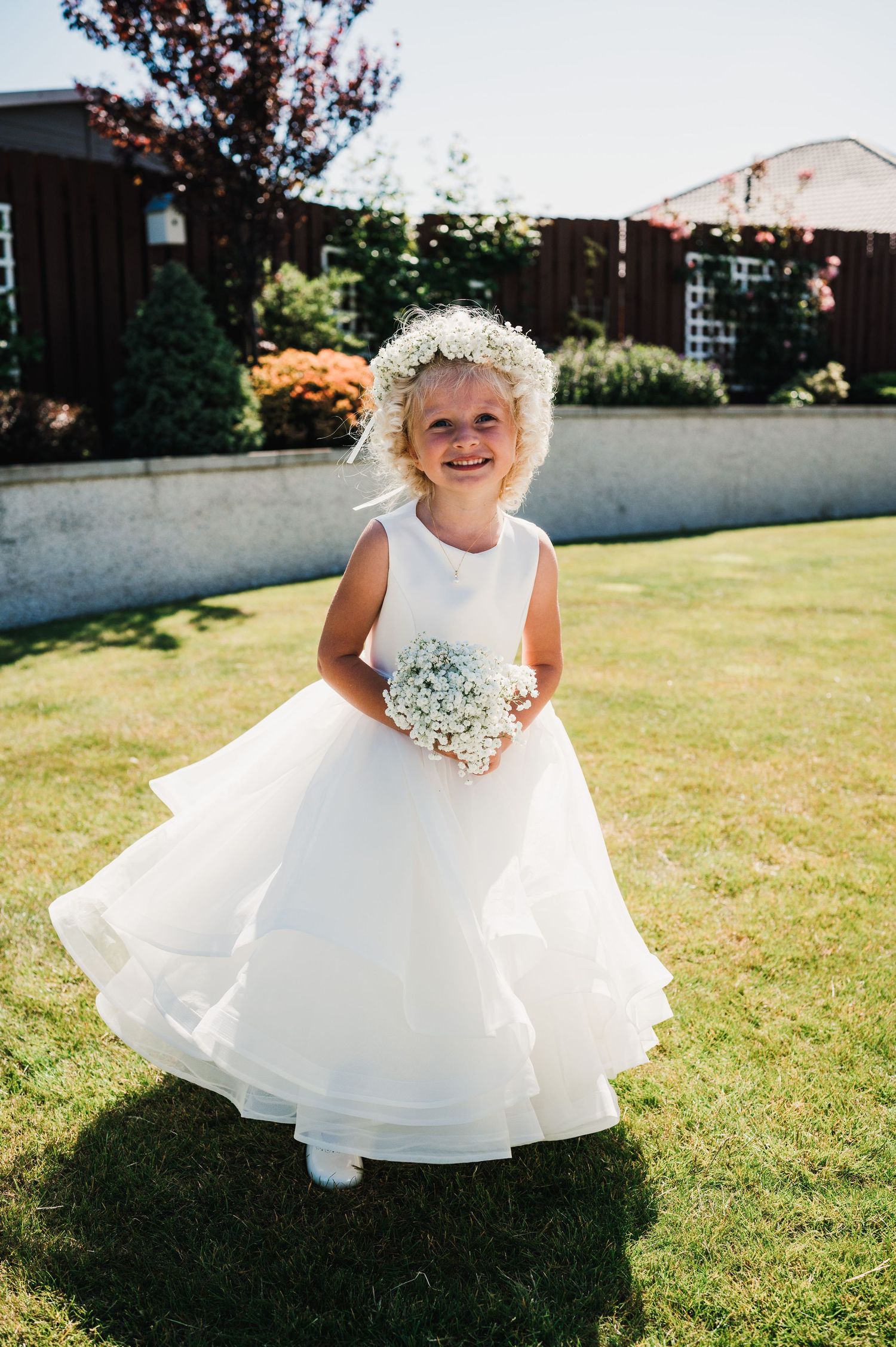 A flower girl in a white dress and floral crown twirls in a sunny garden.