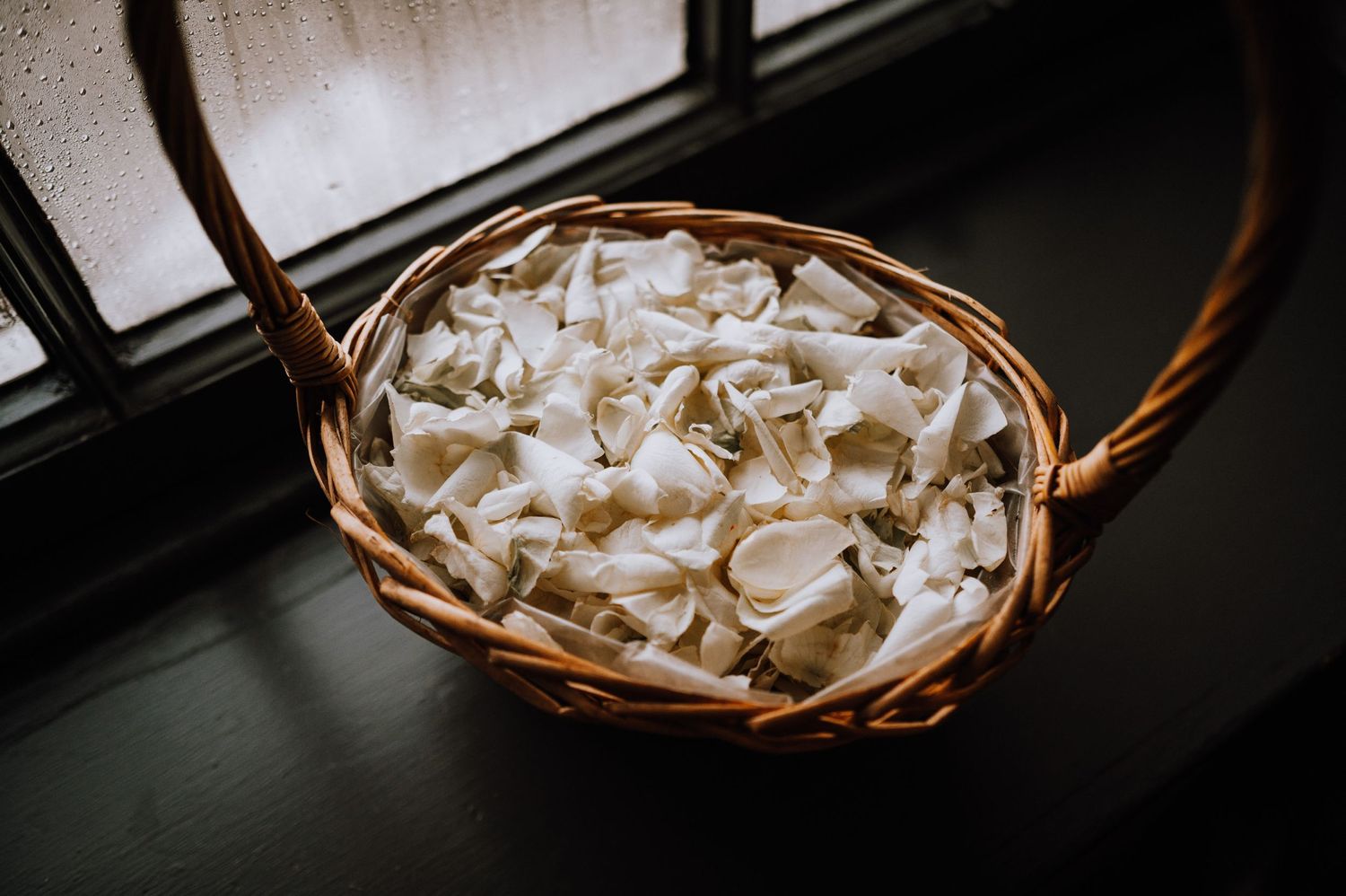 White flower petals arranged in a rustic woven basket sitting by a window.