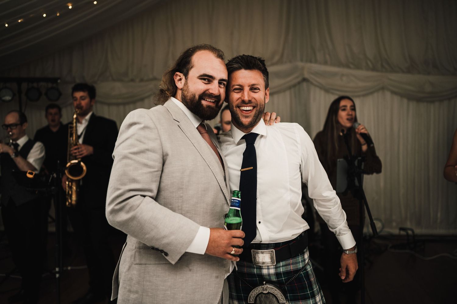 Two wedding guests enjoying drinks and laughter at a reception with traditional Scottish attire.
