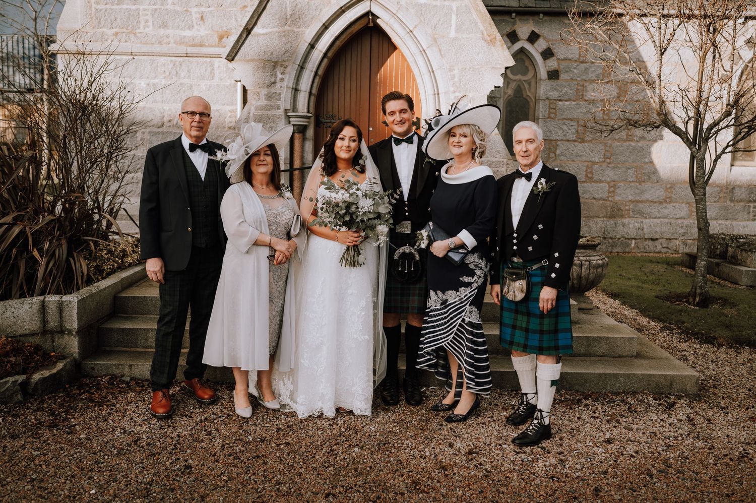 Family group photo outside church with bride in white and Scottish traditional dress elements.