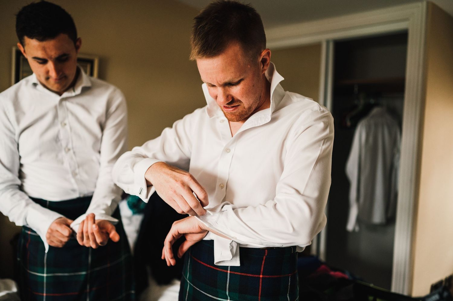 Two people in white shirts adjust traditional Scottish kilts while getting dressed.