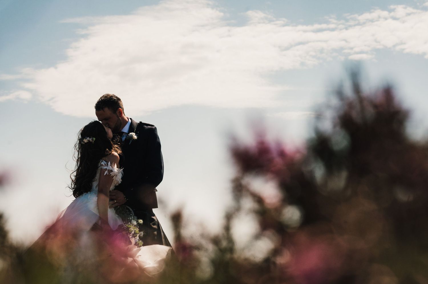 Artistic wedding photo with soft focus flowers in foreground against blue sky.