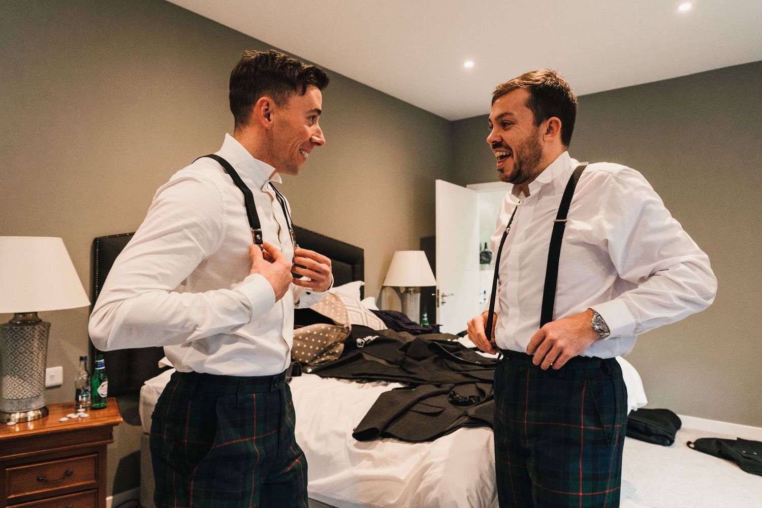 Two groomsmen in white shirts and suspenders share a laugh while getting ready.