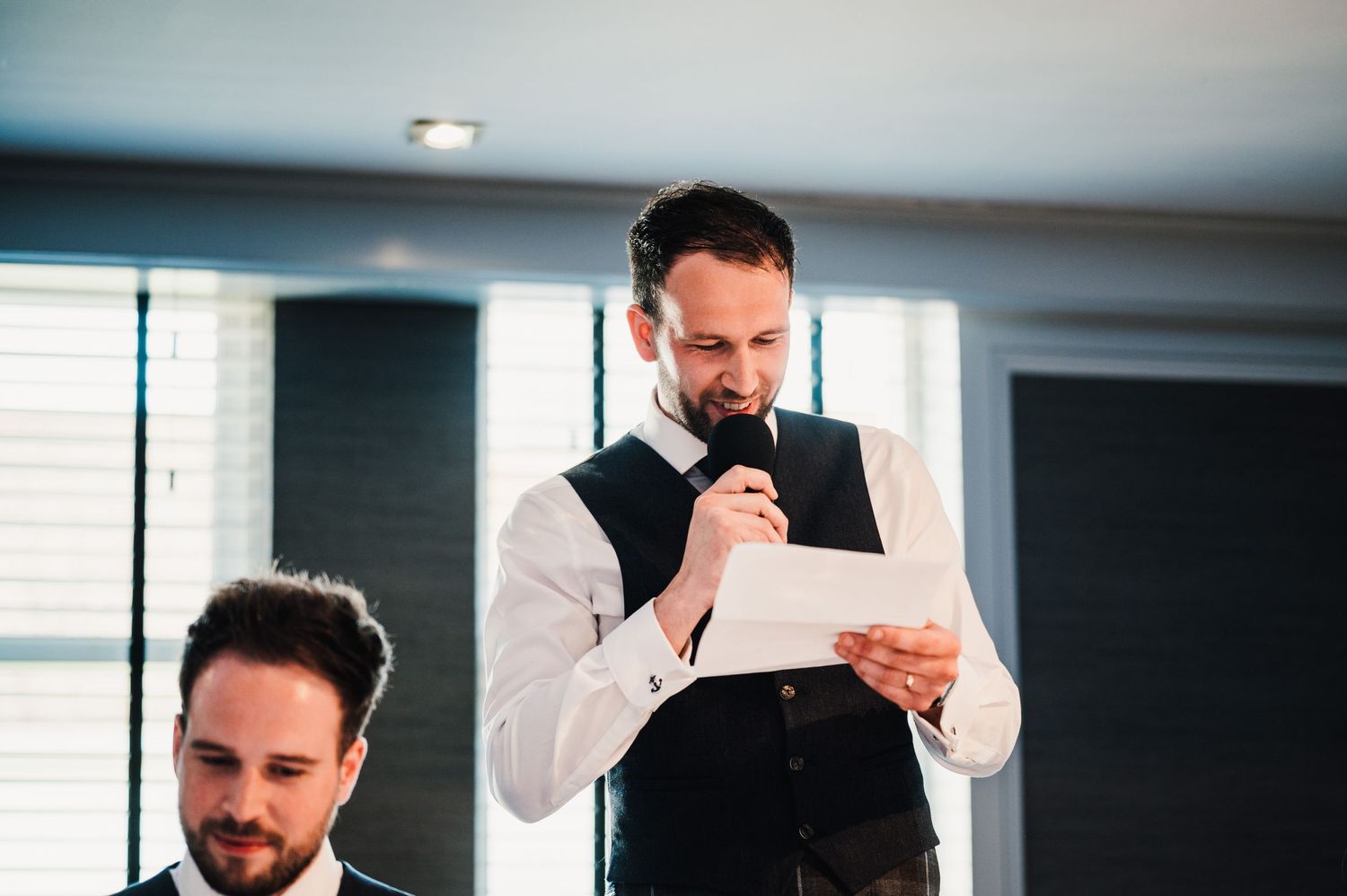 A wedding guest in formal attire delivers a speech from a paper at a reception.