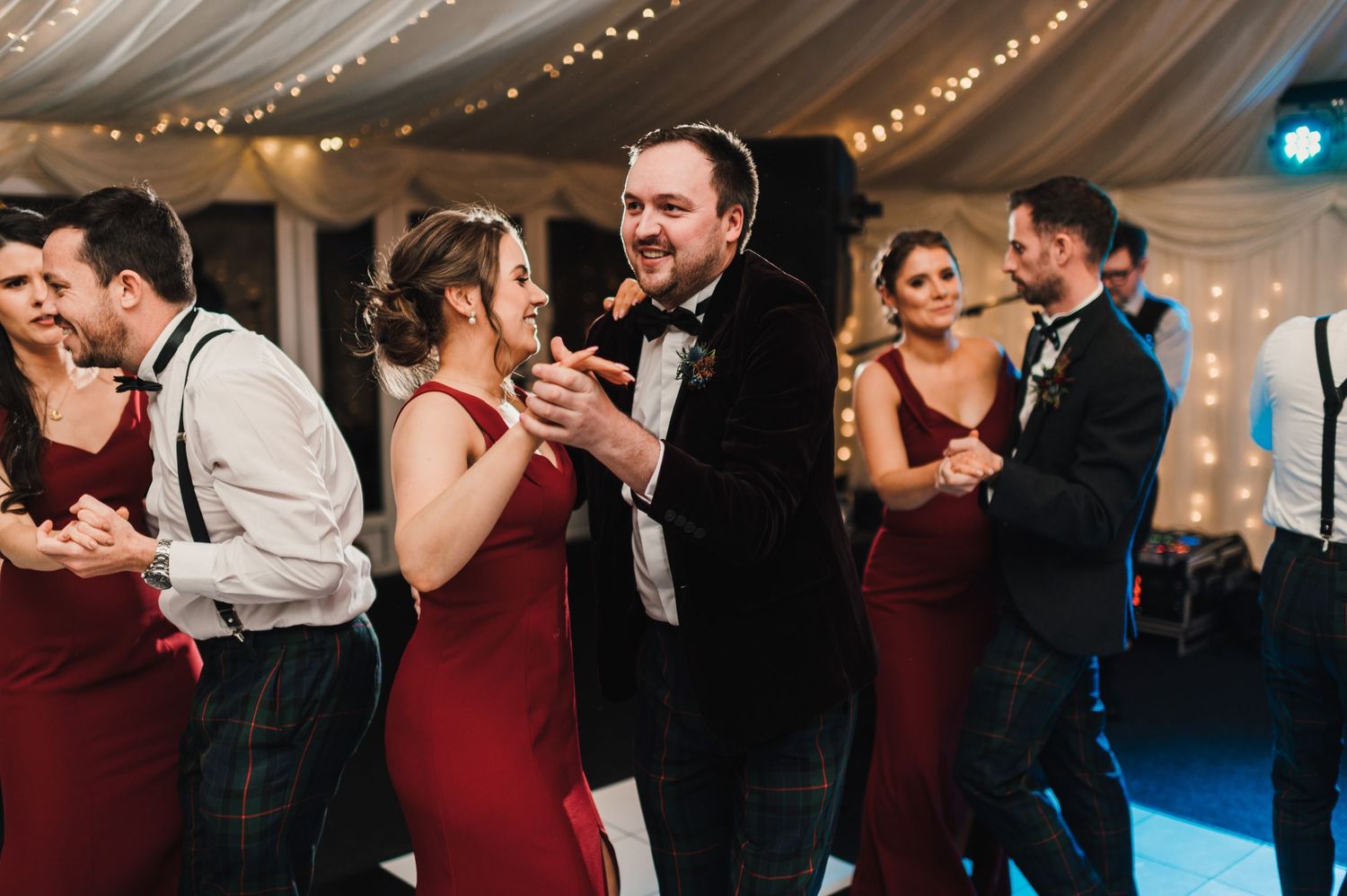 Wedding guests dance energetically in formal attire at a reception under twinkling lights and draped fabric decor.