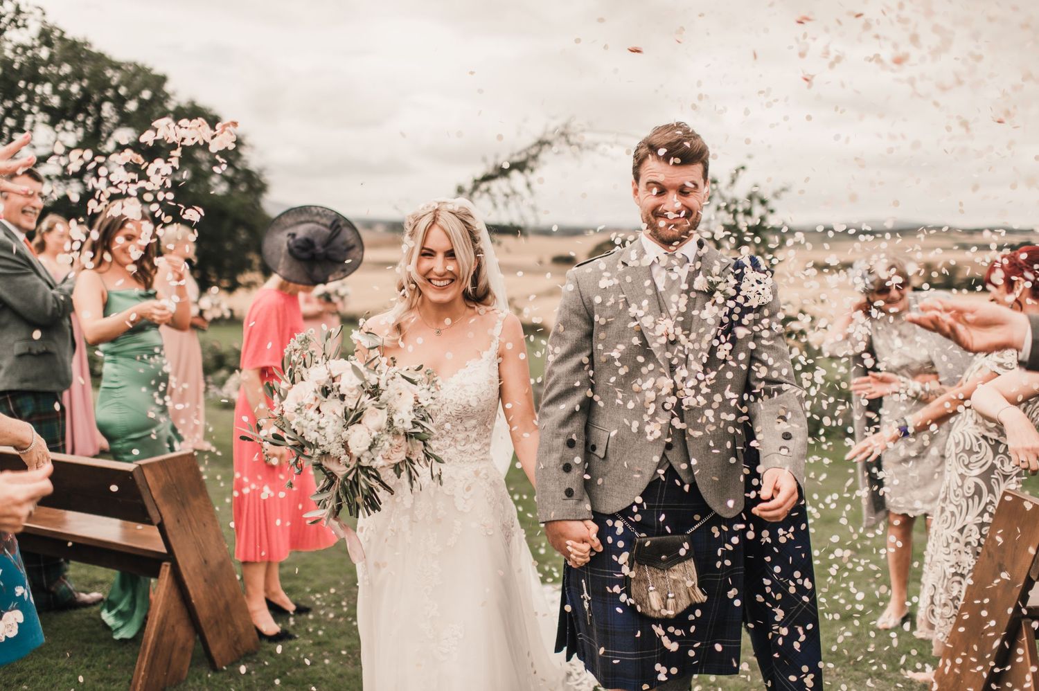 Wedding guests shower the couple with confetti on a hilltop overlooking pastoral fields.
