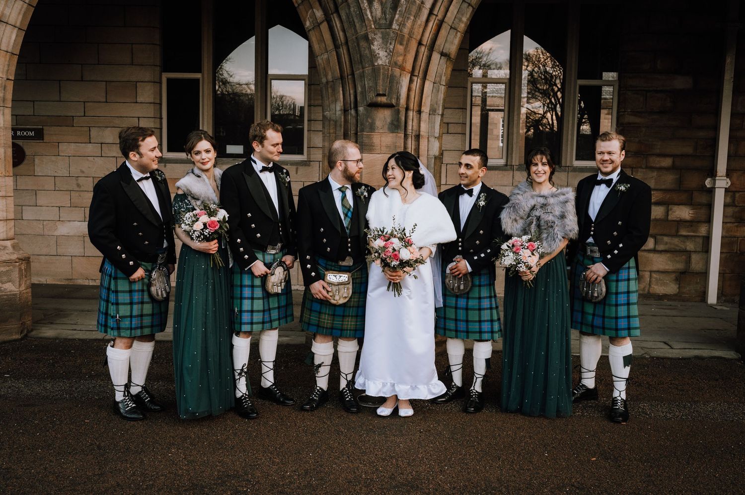 A wedding group wearing traditional Scottish attire poses under stone church archways.