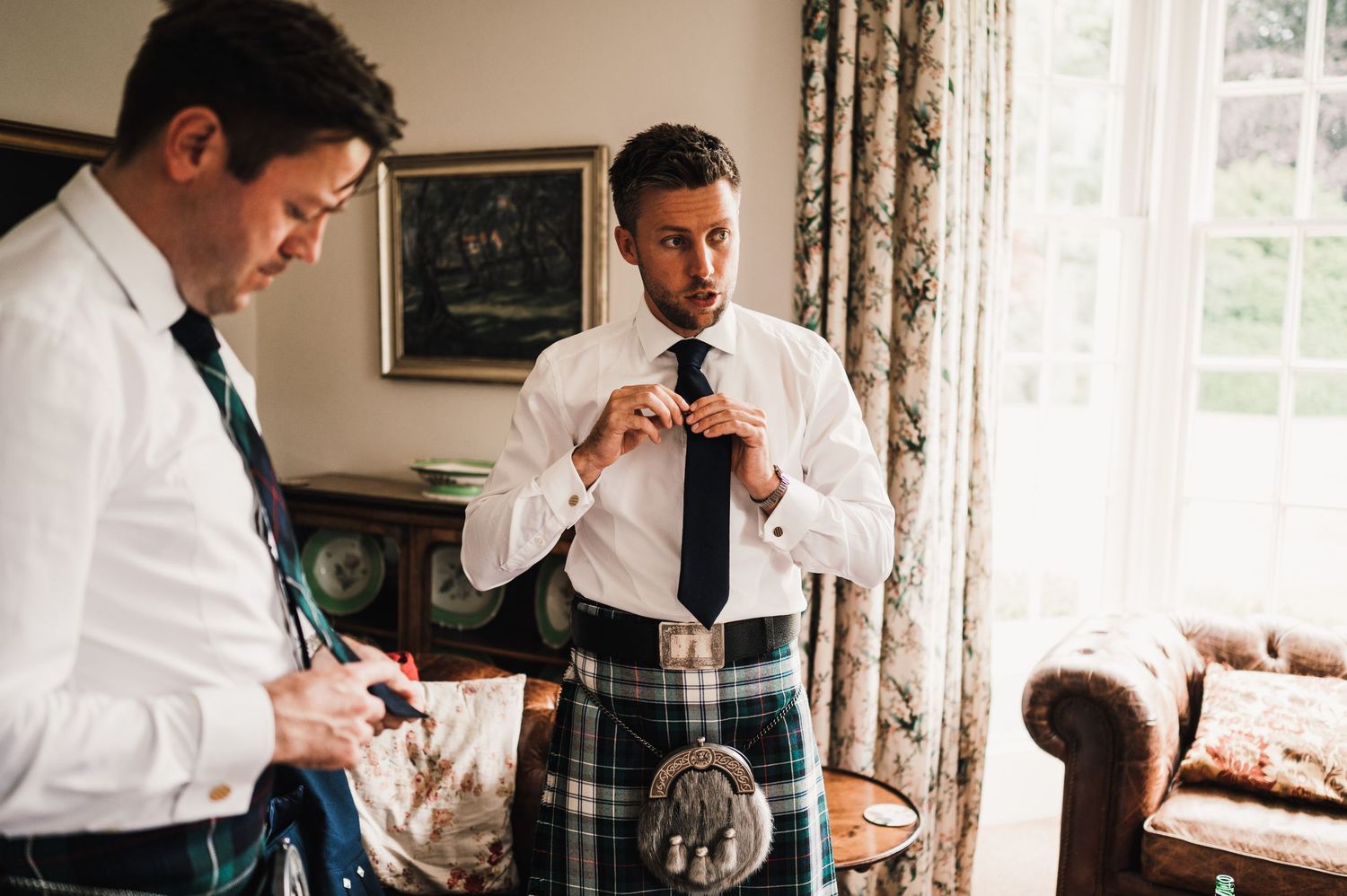 Men in formal Scottish attire preparing for an event by a window.