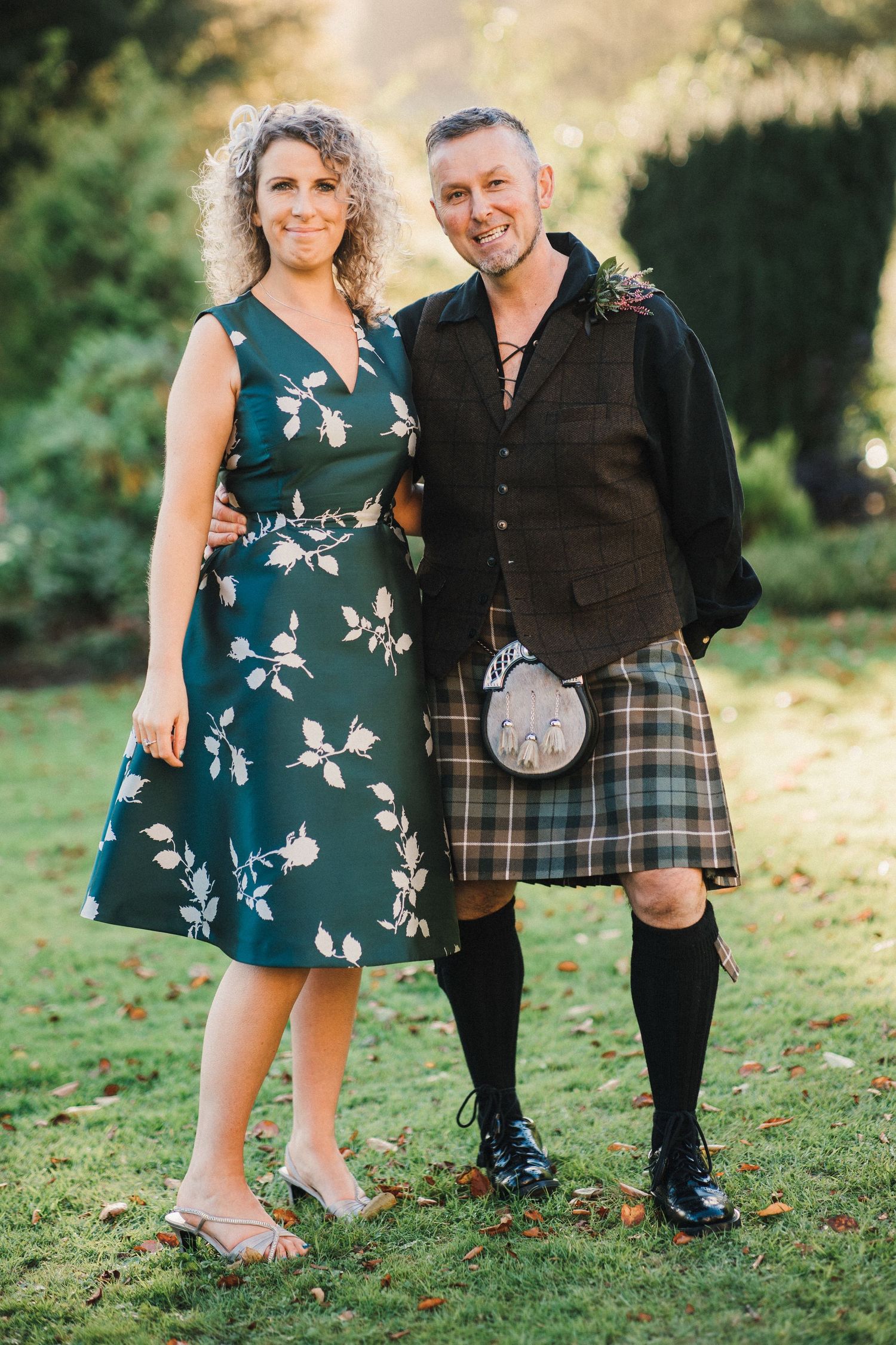 A couple poses on grass, with the man wearing traditional Scottish kilt and the woman in a floral green dress.