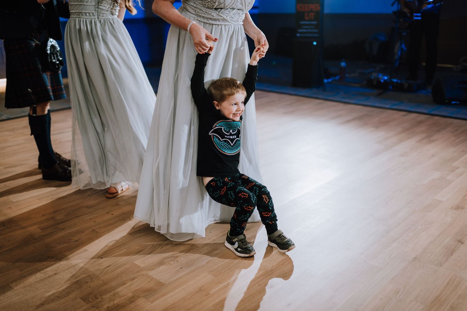 A young child in casual clothes playfully dances between formal dresses at a wedding reception.