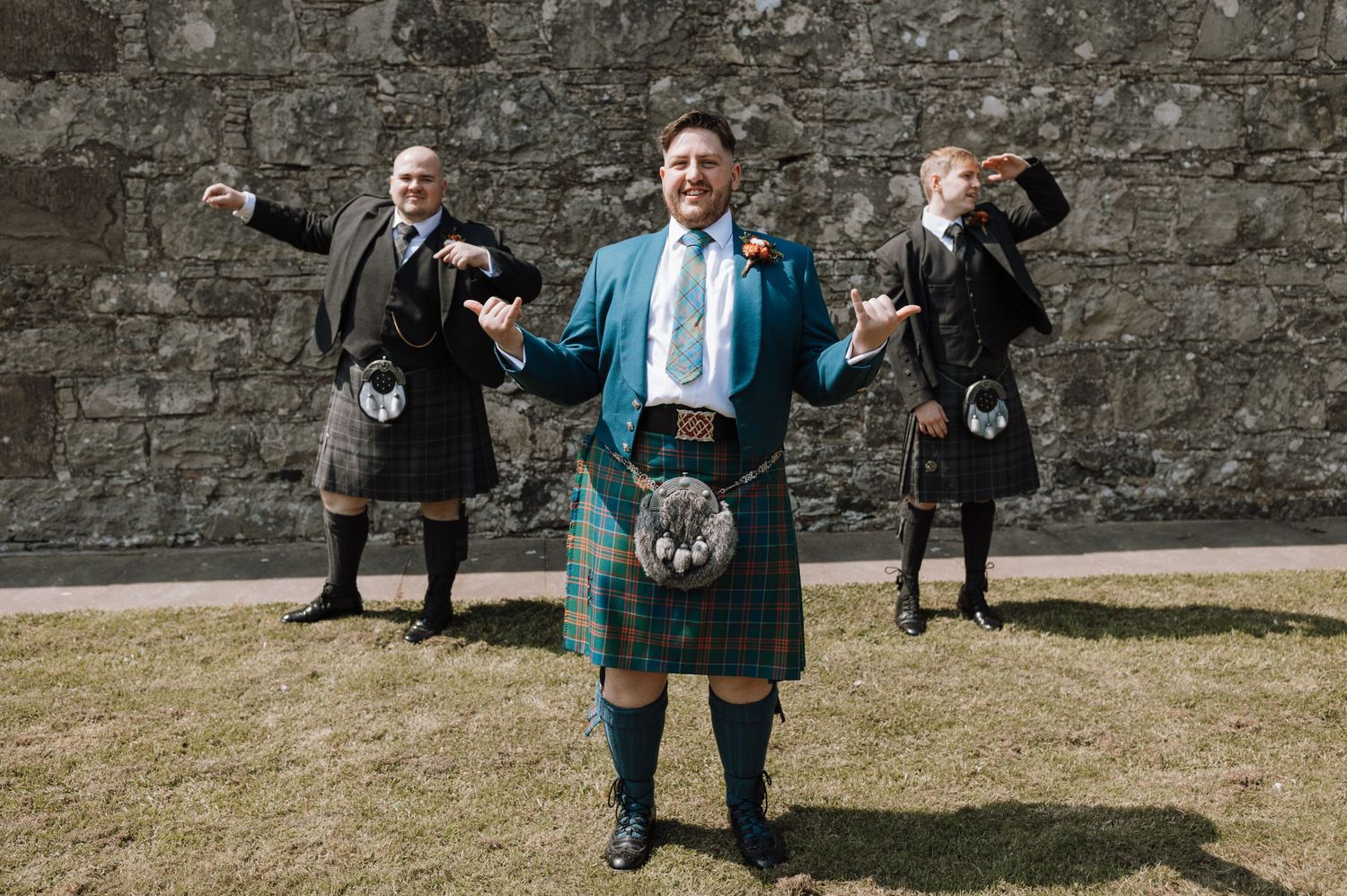 Three people in traditional Scottish kilts pose playfully against a stone wall.