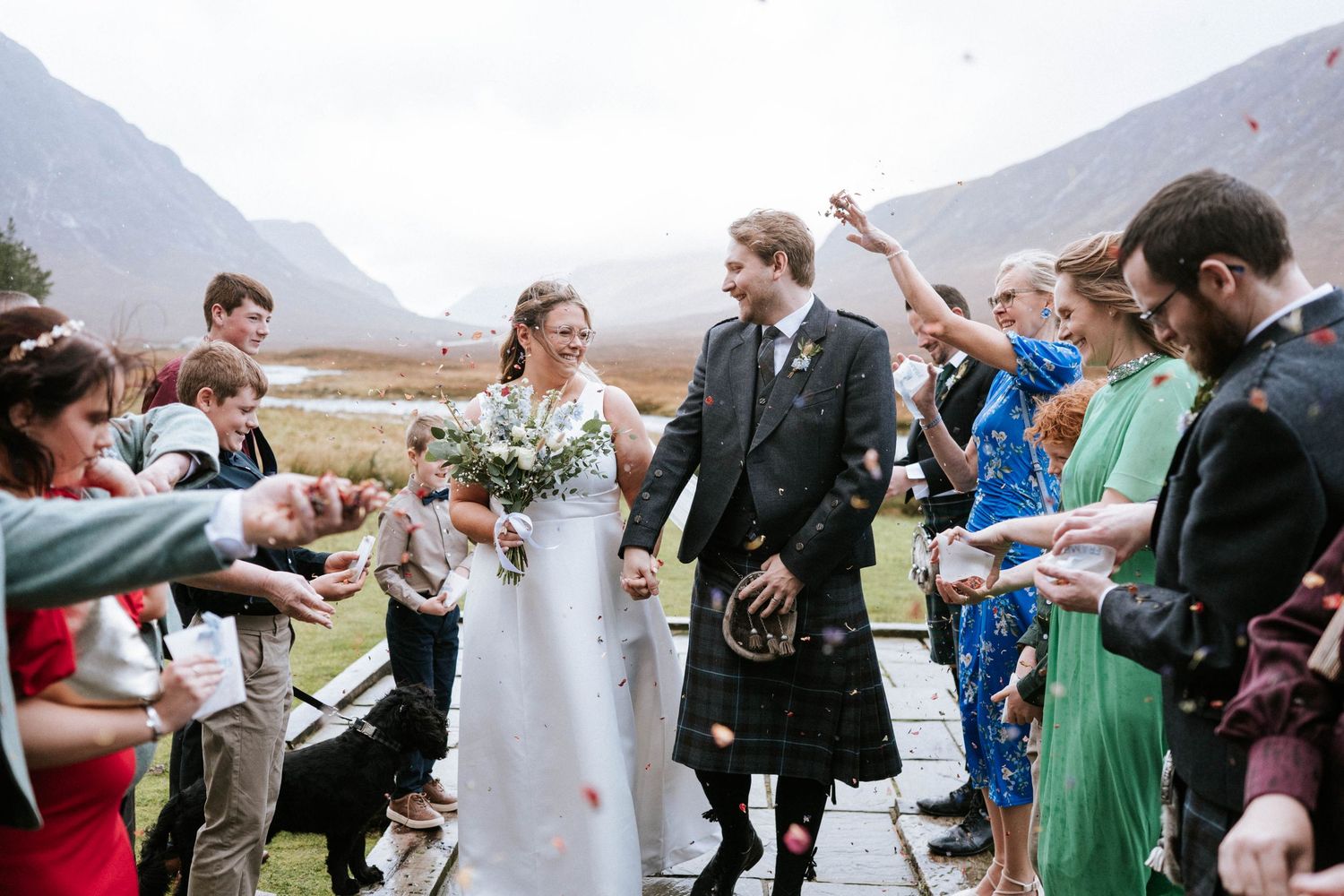 Wedding guests gather around a white tiered cake at an outdoor celebration with mountains in the background.