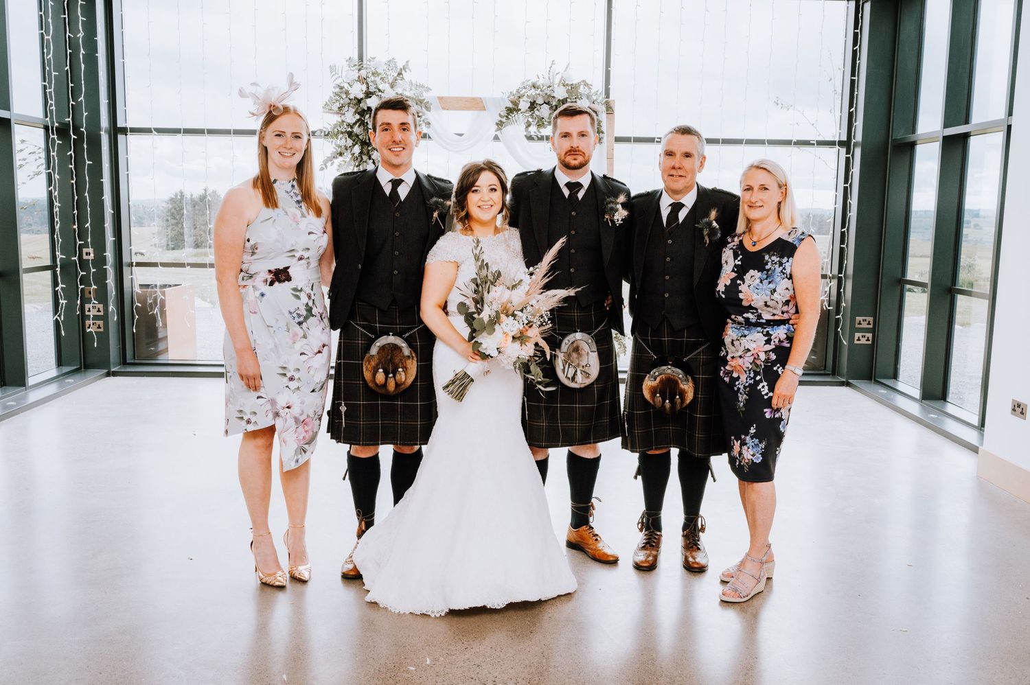 Wedding party members pose in mix of kilts and formal attire in bright modern setting.