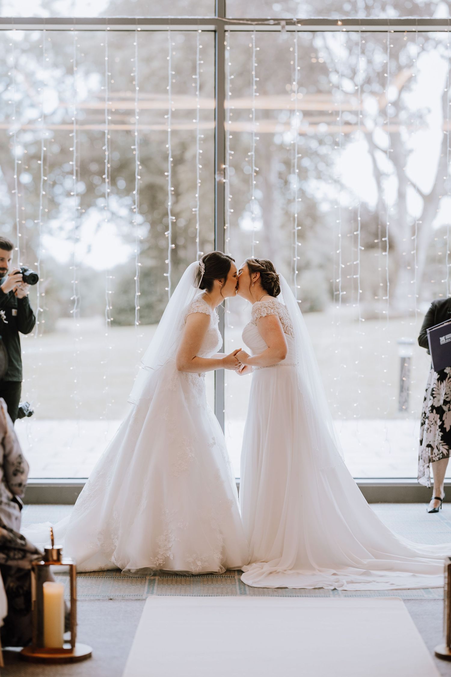 Two brides share an intimate moment under hanging fairy lights beside a large window on their wedding day.