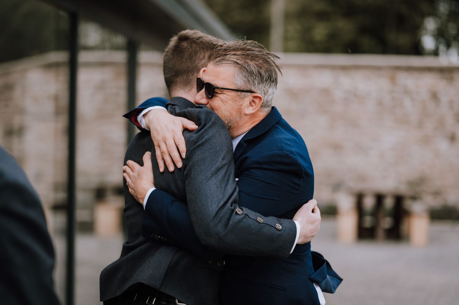 Two men in formal attire share warm hug during outdoor celebration.