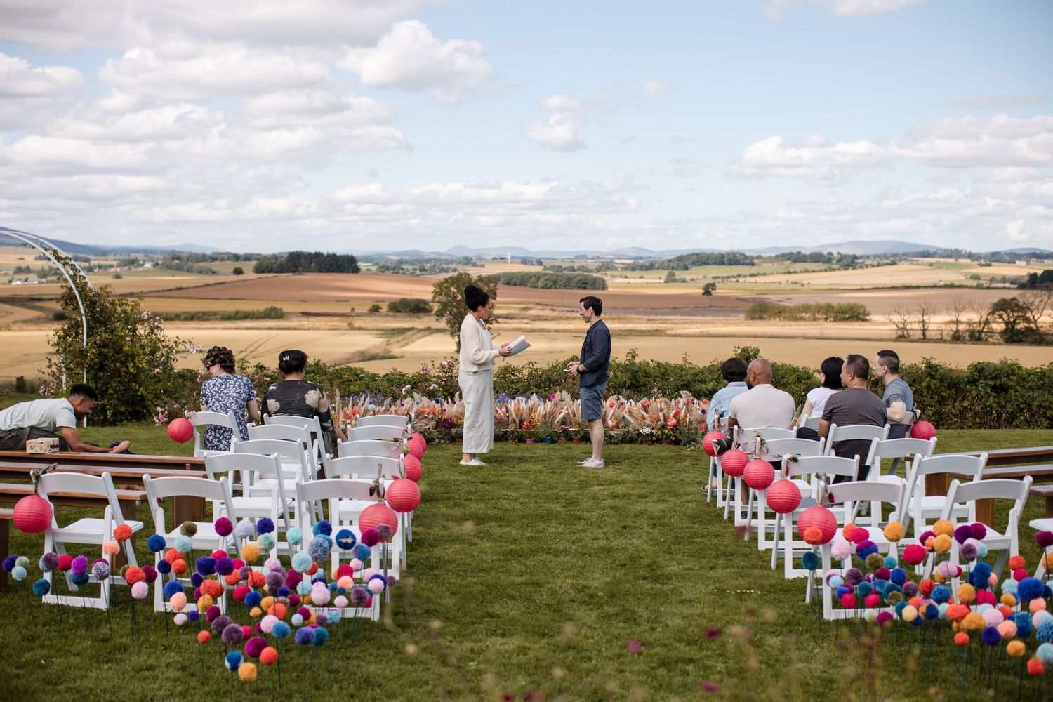 Outdoor wedding ceremony setup with white chairs and colorful balloon decorations against a rural landscape.