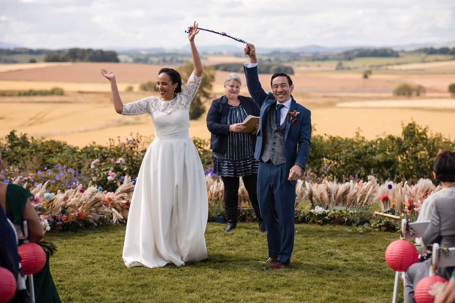 Wedding guests celebrate outdoors with raised glasses against a scenic countryside backdrop during sunset.