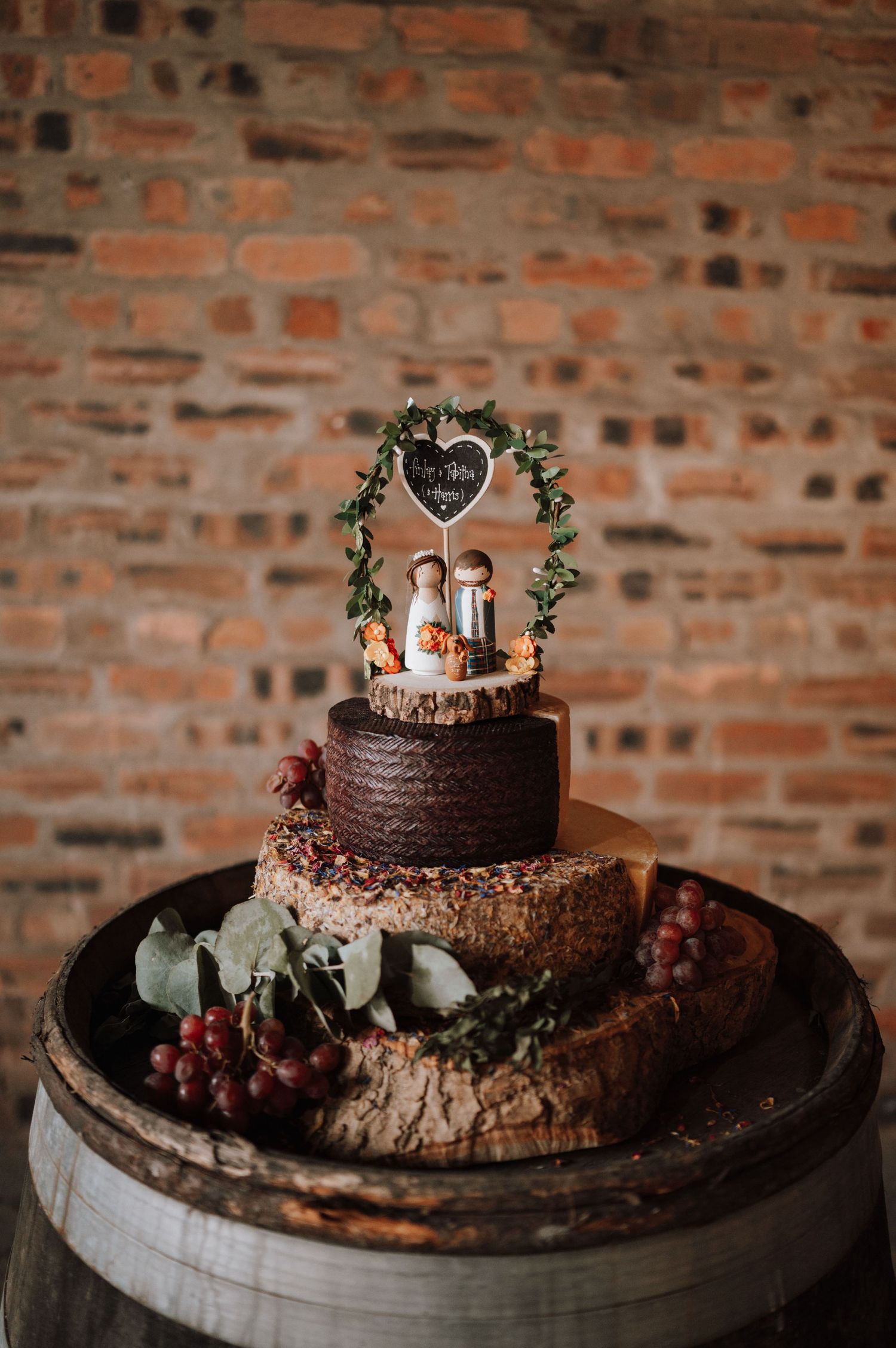 Two-tier chocolate wedding cake with greenery and grapes displayed on wooden barrel.