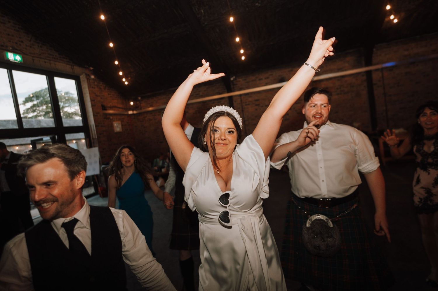 Wedding guests dancing joyfully at an indoor reception with string lights and exposed brick walls.