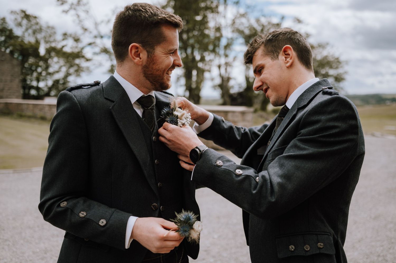 Two men in formal Scottish attire help each other with boutonniere adjustments.