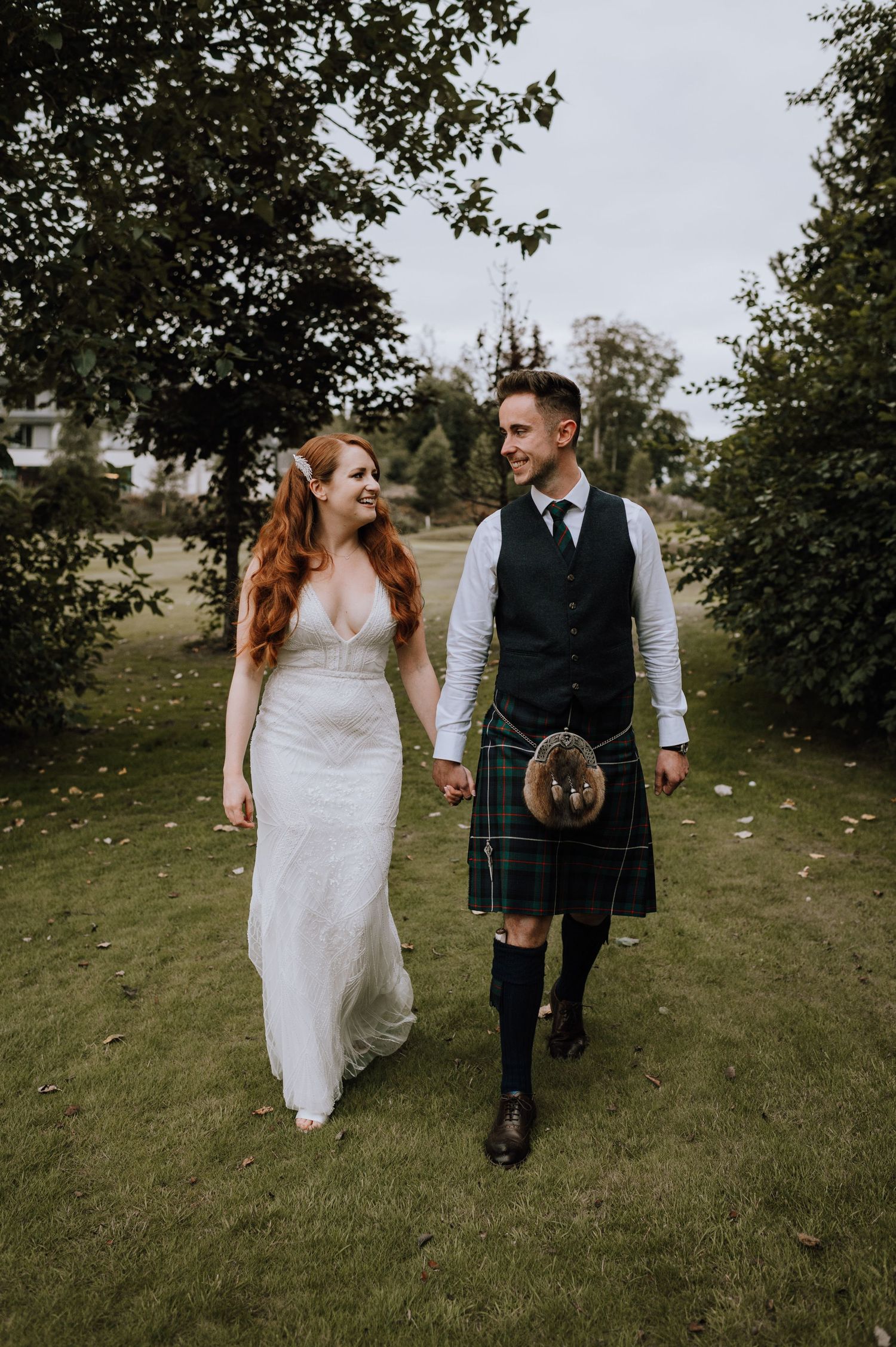 A bride in white dress and groom in traditional kilt walk together on grass.