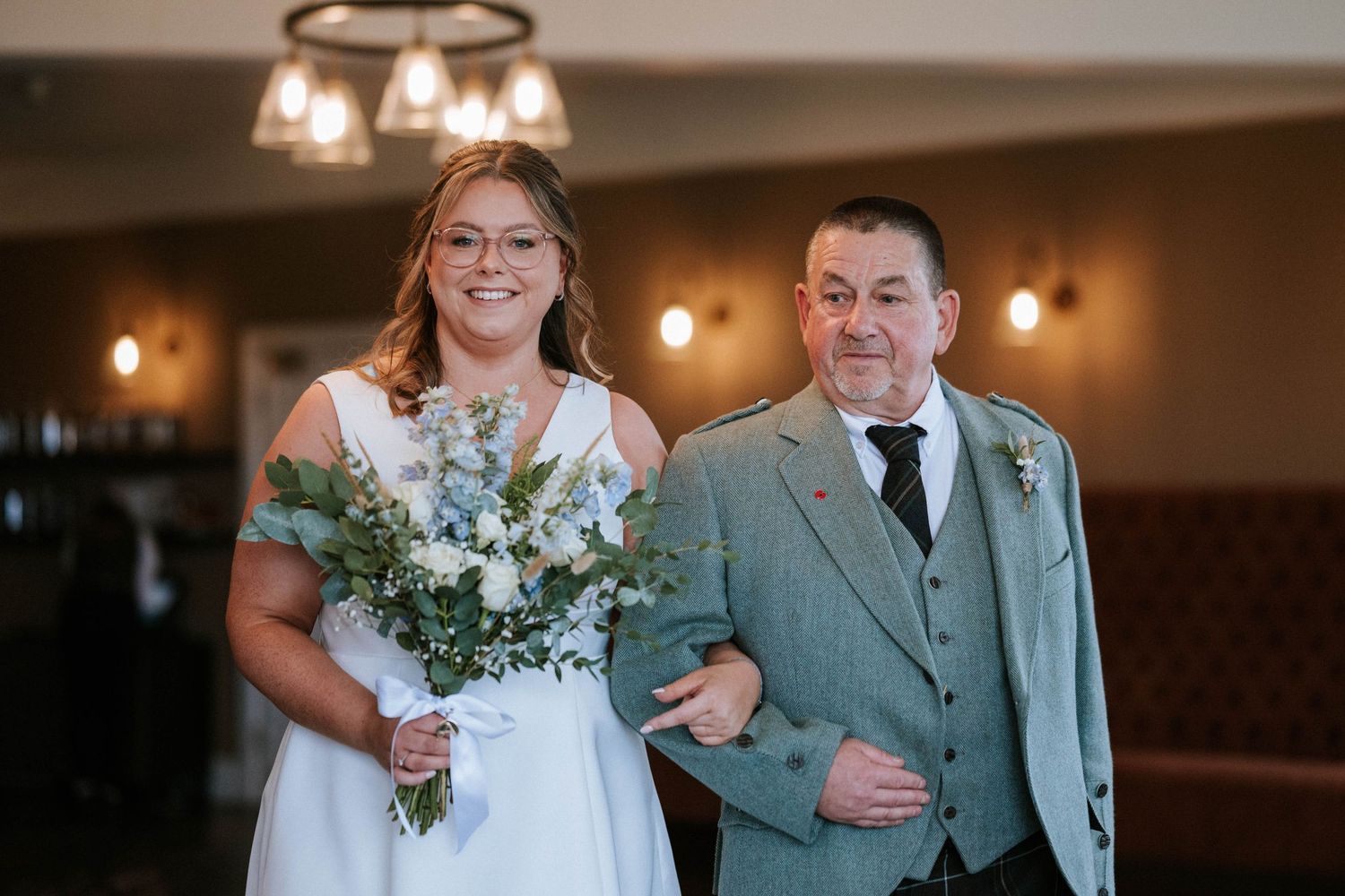 A bride with a bouquet and companion in gray suit walk down an elegant hallway.