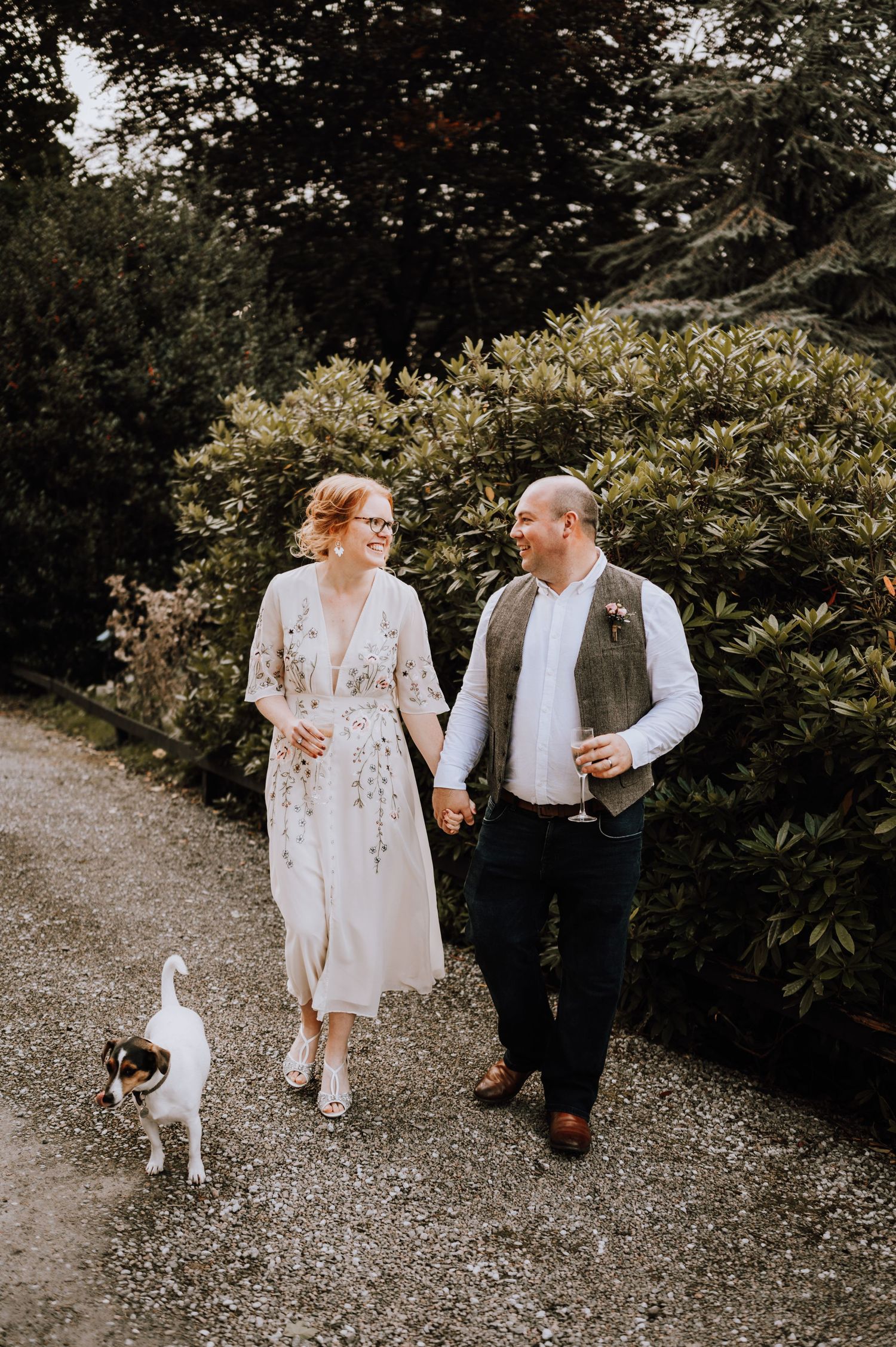 Couple walking on gravel path with small dog through lush garden setting.