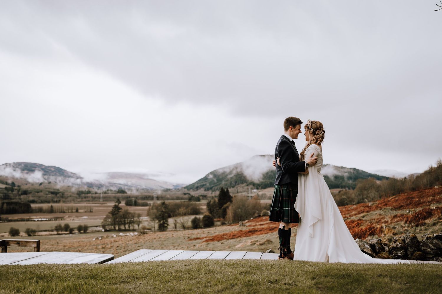 Couple stands on Scottish hillside in traditional wedding attire with mountainous backdrop.