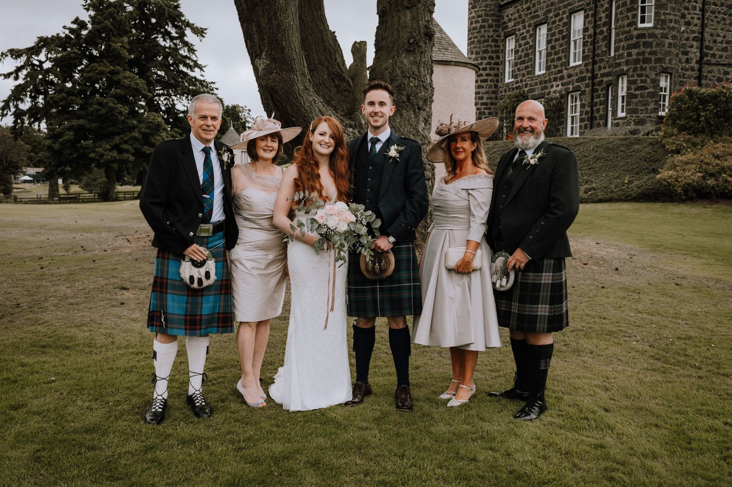 Small group in Scottish wedding attire including kilts pose together on castle grounds.