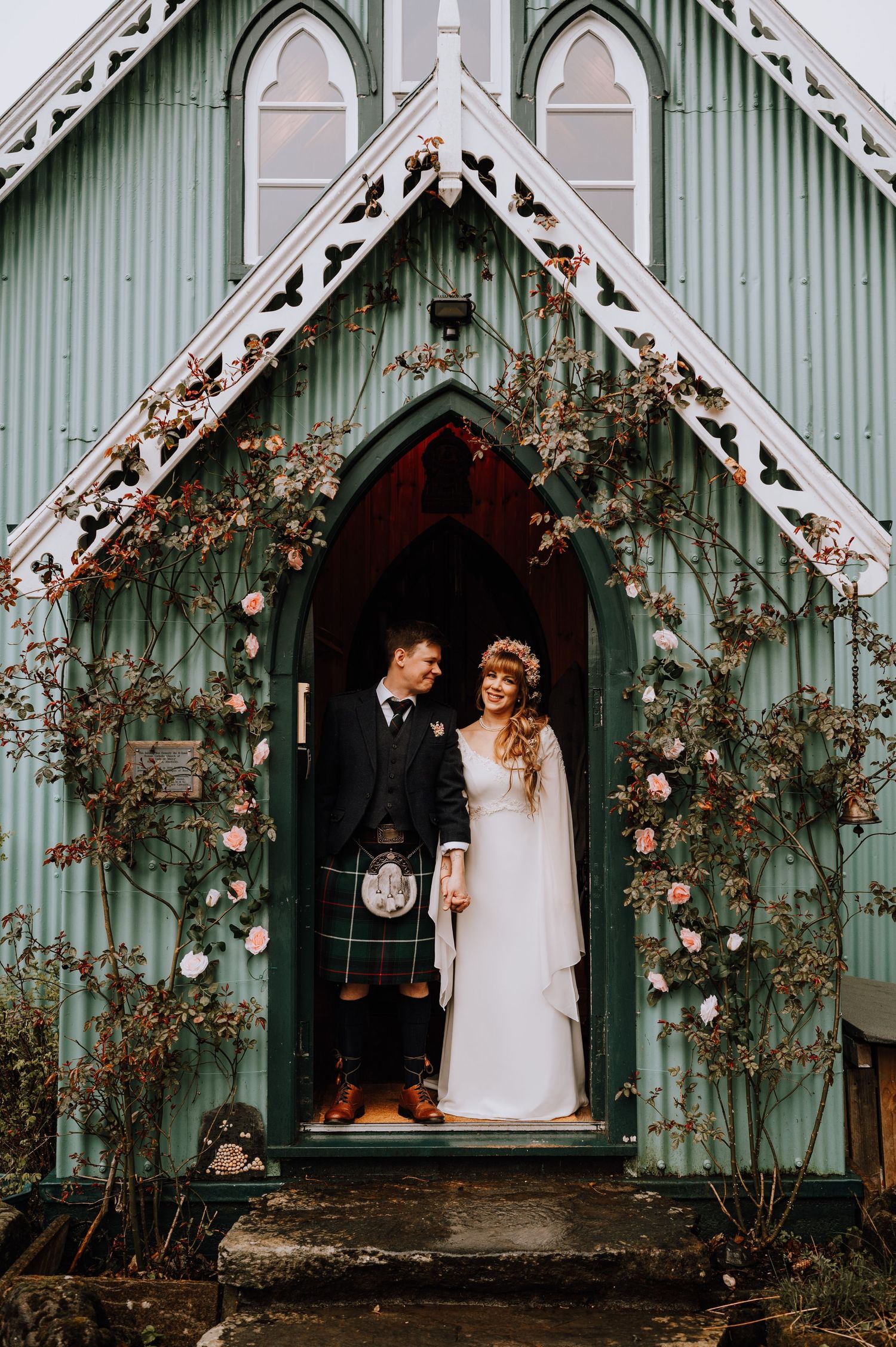 Wedding couple stands in doorway of mint green chapel decorated with climbing roses.