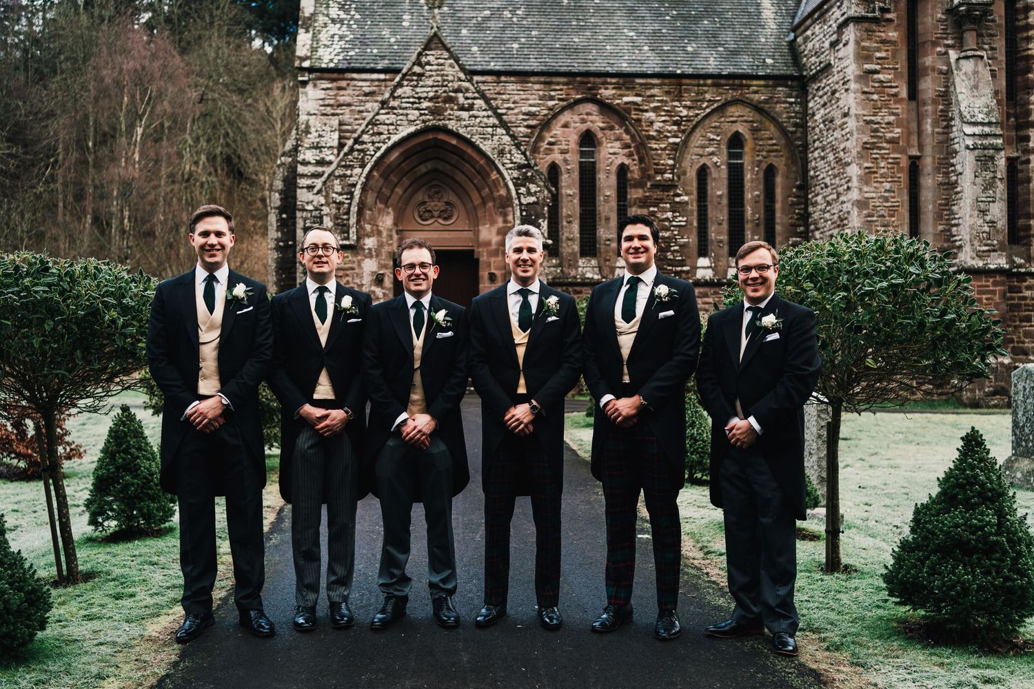 Groomsmen in formal black suits pose together outside a gothic stone church.