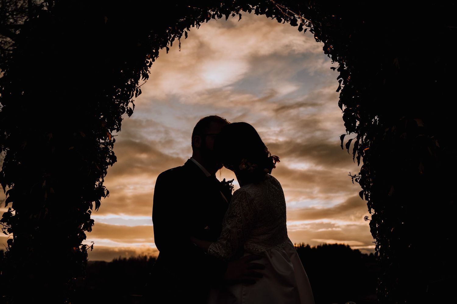A silhouetted couple shares a tender moment under an arch against a dramatic sunset sky.