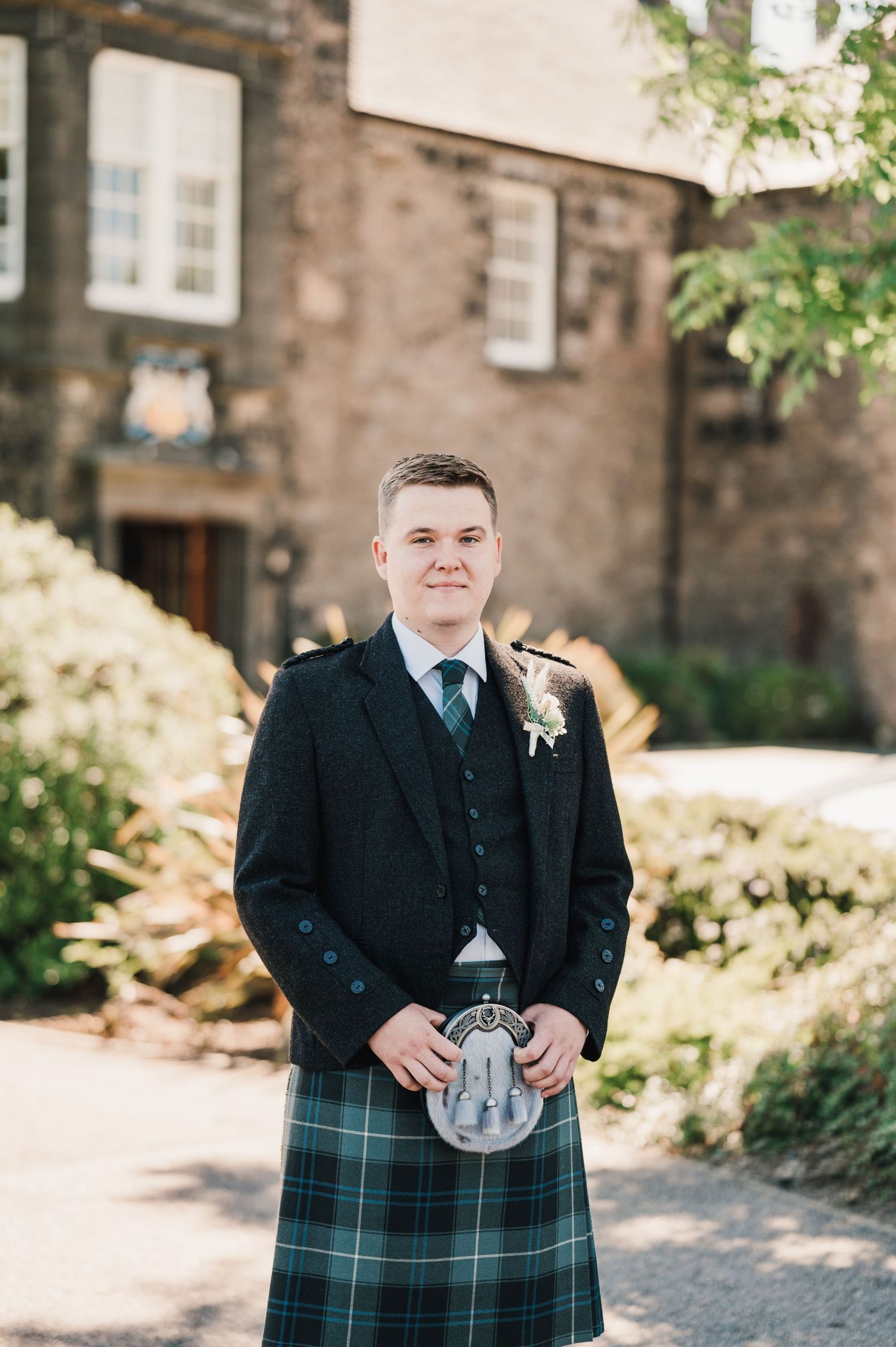 A person in Scottish formal wear stands outside a stone building on a sunny day.