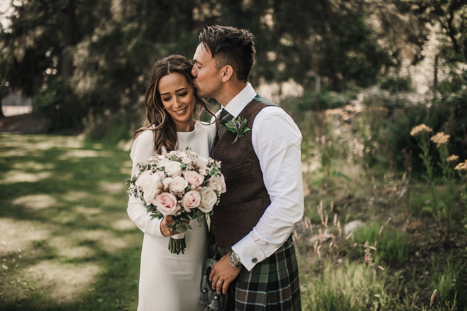 Intimate wedding moment captured in forest setting with Scottish-themed attire and pink bouquet.
